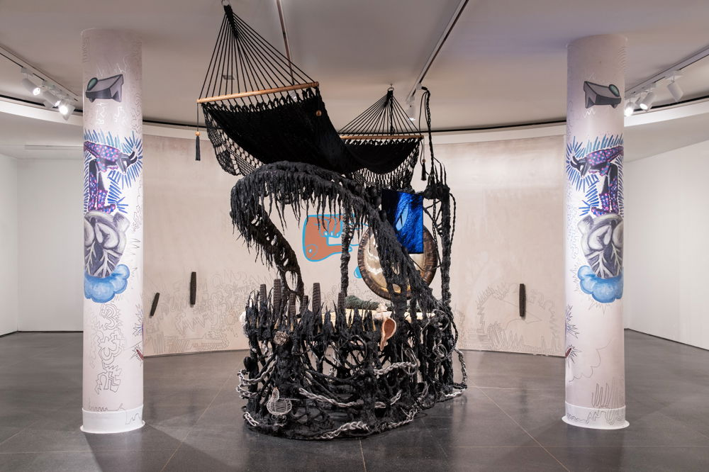 A gallery installation features a large abstract sculpture centered between two pillars. The sculpture is made of black fibers and a gong and arches out like the jawbone of a whale. A hammock suspended from the ceiling drapes over the top of the structure. The pillars are each painted with abstract designs centering on imagery that resembles an anatomical heart.
