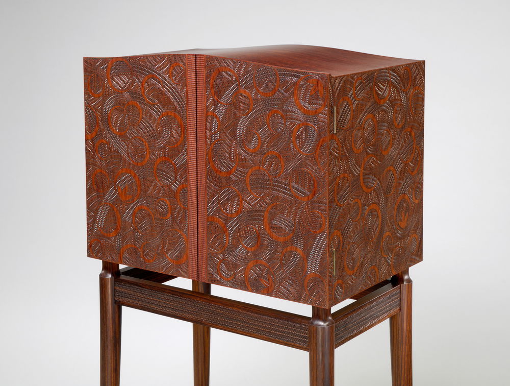 A three-quarter view of a compact two-door rectangular cabinet resting on a stand. The cabinet has carved interlaying circular designs that look like red eclipses over toothy gray patterned spirals.