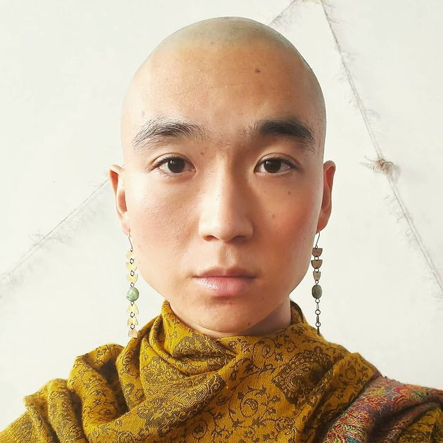 Eddy, a trans non-binary artist of Korean descent, has a shaved head and is wearing long, dangling earrings and a deep yellow scarf. They have dark eyes and are looking into the camera. In the background, frayed string forms a loose triangle on the wall.