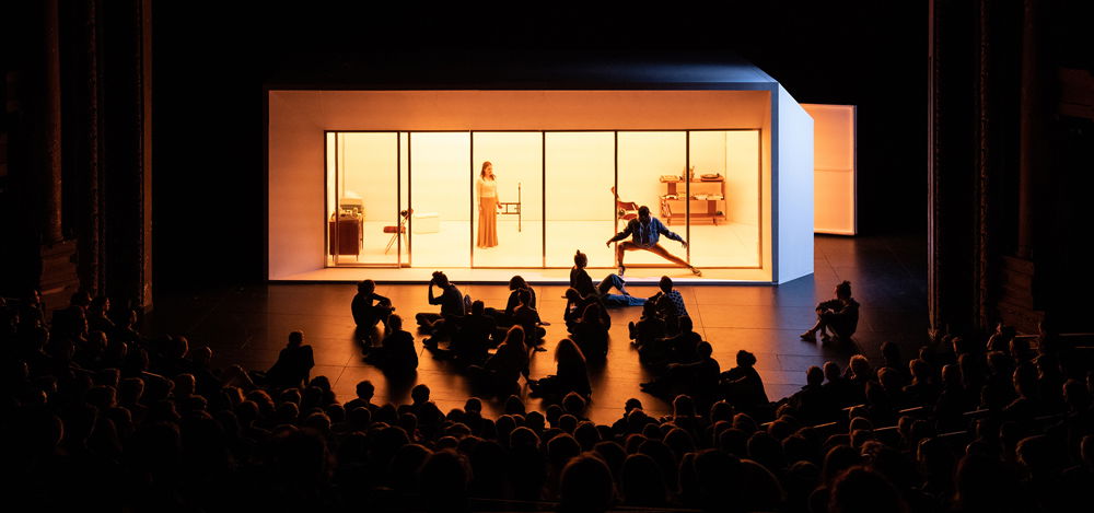 A photograph of a stage, mid-performance. The set is a long rectangular structure with many glass doors, lit from the inside to reveal furniture and a woman standing alone. In front of the windows, one person dances in silhouette while other figures lounge downstage.