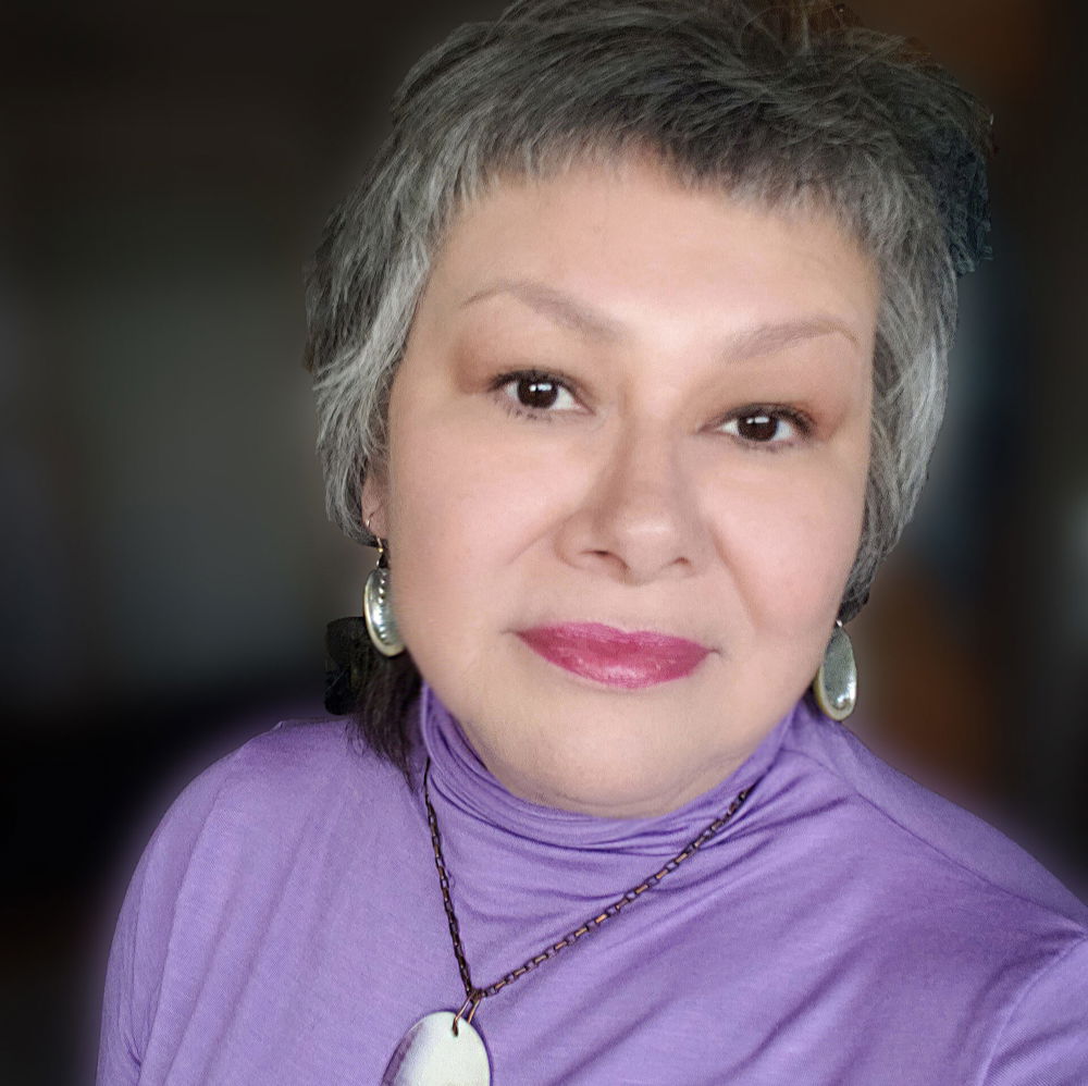 Delina, a woman with short hair and pink lipstick, smiles slightly. She is wearing a purple turtleneck, dangling earrings, and a pendant necklace.