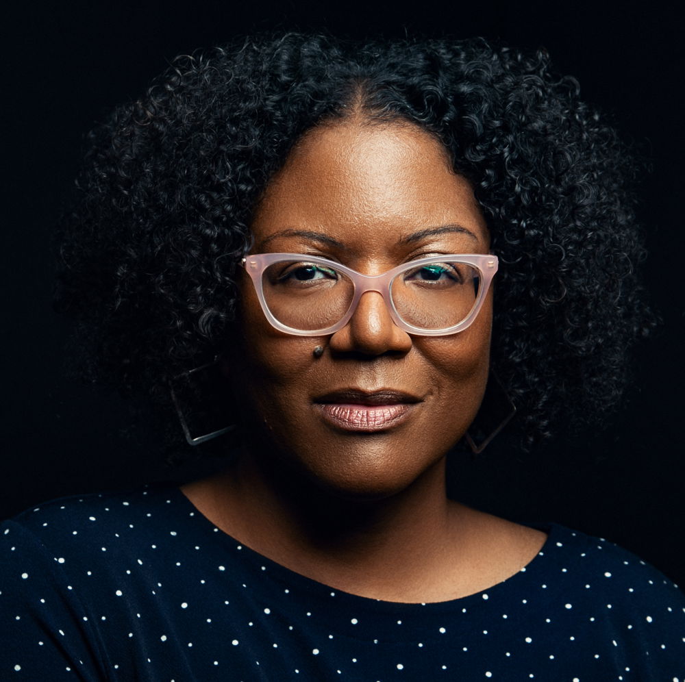 Honorée, a milk chocolate-brown Black woman with chin-length, curly black hair, wears light pink glasses, geometric earrings, and a navy dress with tiny white polka dots.