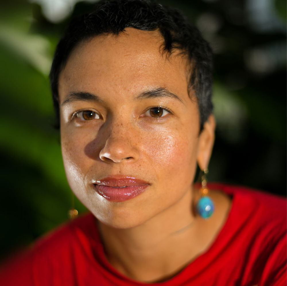 A person with brown skin, brown eyes, and short black hair, looks into the camera. They are wearing a red shirt, one blue earring, and have small tattoos on their neck.