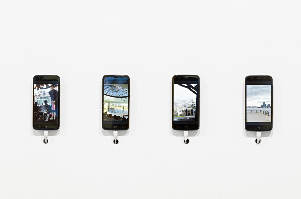 Four iPhones mounted on a white wall display photos: a person in a cafe, at a public pool, in the rain holding an umbrella under a tree, on a pier.