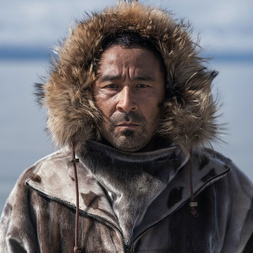 A portrait of a man in a seal skin jacket on the shores of Aak'w Kwaan Territory. His face is stoic, as he looks directly ahead. Under his fur hood is a tuft of black hair peeking out, and his face has dark stubble around the chin.