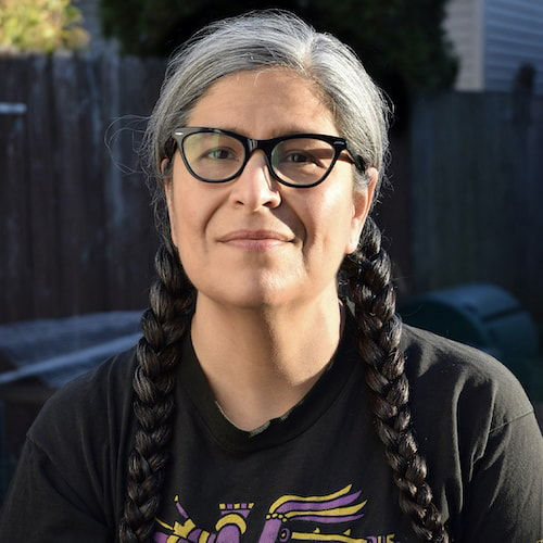 A fifty-year-old Latinx person with glasses squints at the viewer. Their salt-and-pepper hair is in two long braids, and they are wearing a t-shirt with Huitzilopochtli on it.