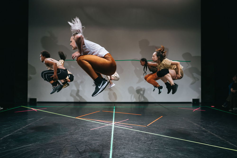 Six performers jump up in perfect unison on an empty stage. The performers are positioned in a circular formation, facing the left with their knees tucked under their torsos and hands at their thighs, resembling the fetal position.