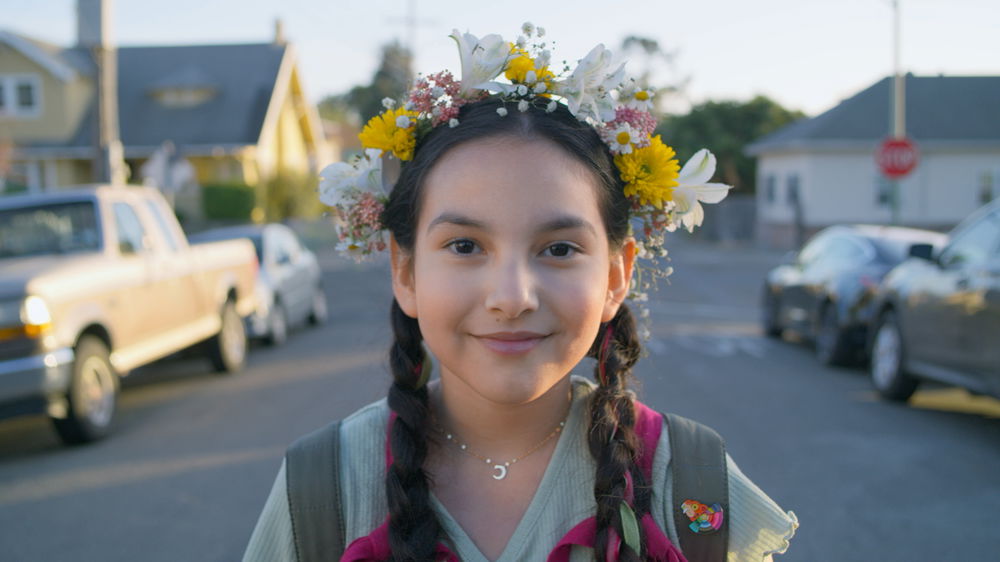 A medium-skinned girl with braided hair and a brightly colored crown of flowers on her head smiles softly at the camera. She stands in the middle of a neighborhood street, where there are parked cars and houses on both sides.