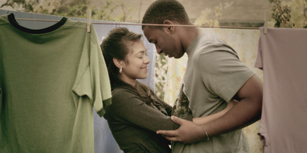 A medium-skinned woman and a medium dark-skinned man stand mid-embrace between outdoor clotheslines draped in various shirts and linens.