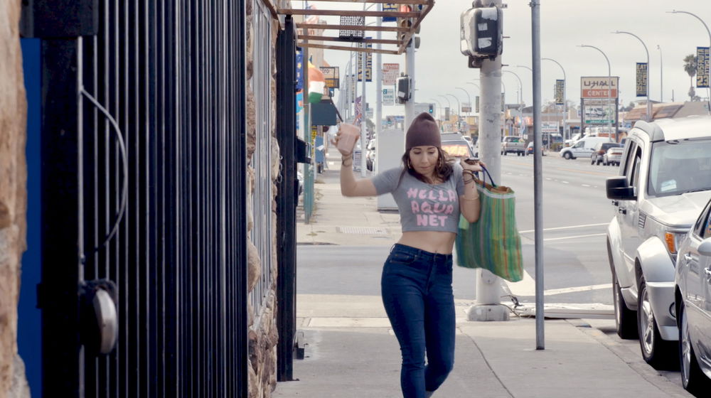 A woman in a gray crop top that reads “HELLA AQUA NET” in pink text and jeans walks down the sidewalk of a busy city street on an overcast day. She is carrying a drink in one hand and a large tote bag in the other, raising them slightly above her body.