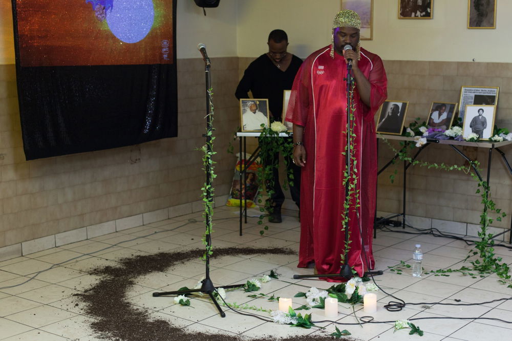 A performer stands behind a microphone stand covered in vines. They wear a gold headdress and long red garment made of a shimmering material. Surrounding the performer is a semi-circle of dirt, flowers, and candles on the floor. Behind the performer is a second person standing by a table covered in photographs of people.