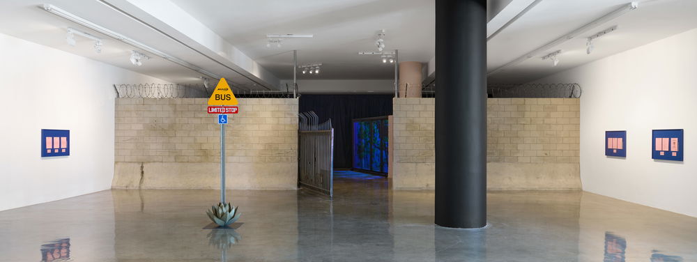 A large gallery space with white walls and a shiny floor is bifurcated by a prison-like cinderblock and barbed wire barrier wall. In front of the wall is a street sign with the words "Bus" and "Limited Stop," and a metal agave plant at its base.