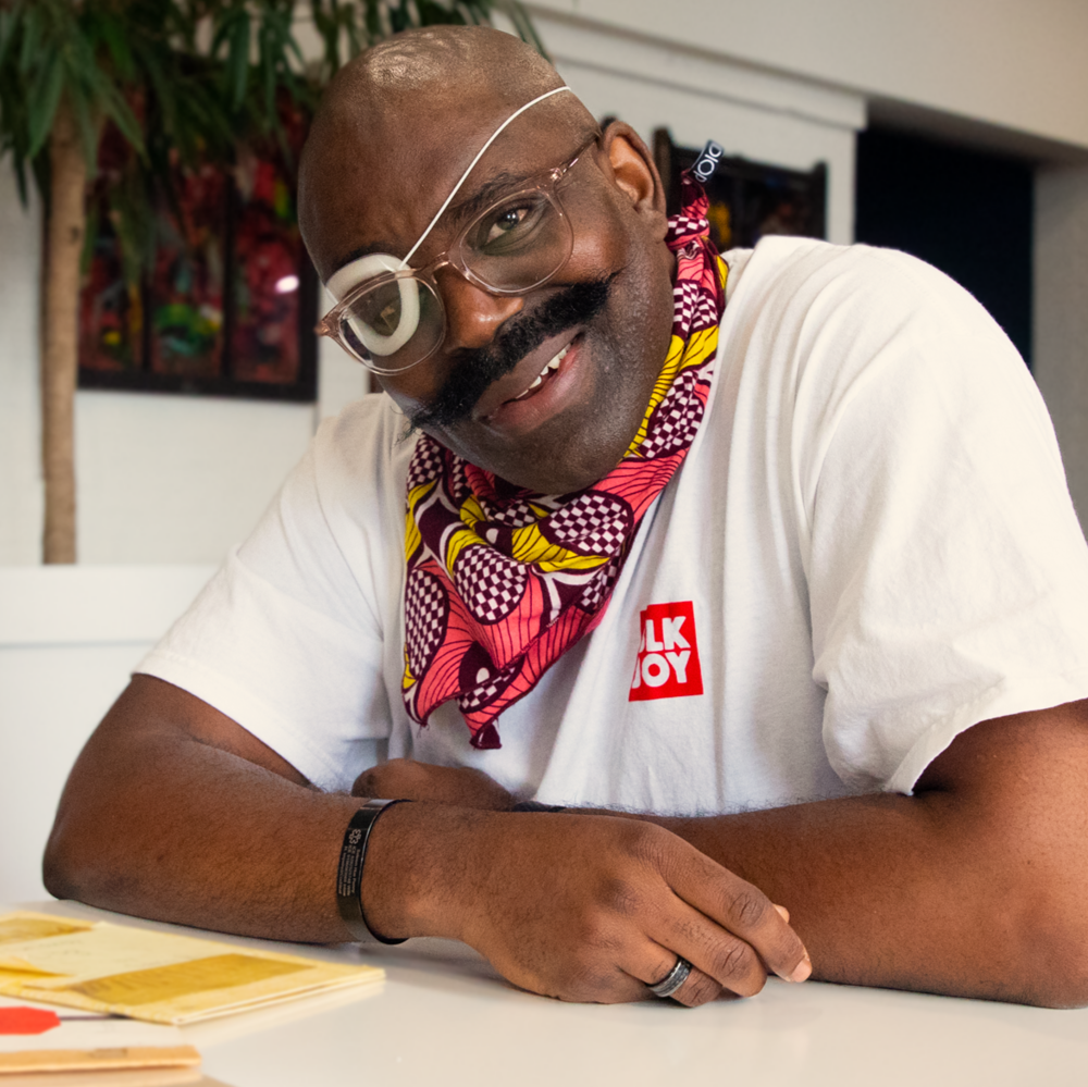 Saleem, a Black man with a partially paralyzed face, an ever-widening smile, and a handlebar mustache, faces the camera. He wears clear-framed glasses, a plastic moisture chamber over his right eye, and a right-sided cochlear implant and sits comfortably hunched over a table with arms loosely folded. He wears a white T-shirt with “Blk joy” printed inside a red square and a pink and yellow floral-print bandana around his neck. Two pamphlet-style books created in first and second grade are on the table.