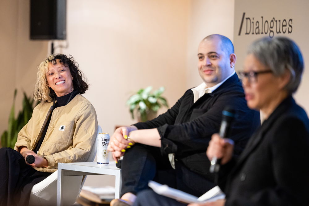 Judilee Reed speaks into a microphone. Seated next to her on the stage, John Paul Morabito and Linda Nguyen Lopez smile warmly.