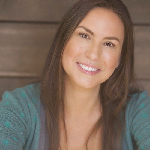 A headshot of a young Cherokee woman with long, straight, brown hair. She leans against a brown wooden wall, smiling out at the viewer. She's wearing a gray shirt with a green cardigan.