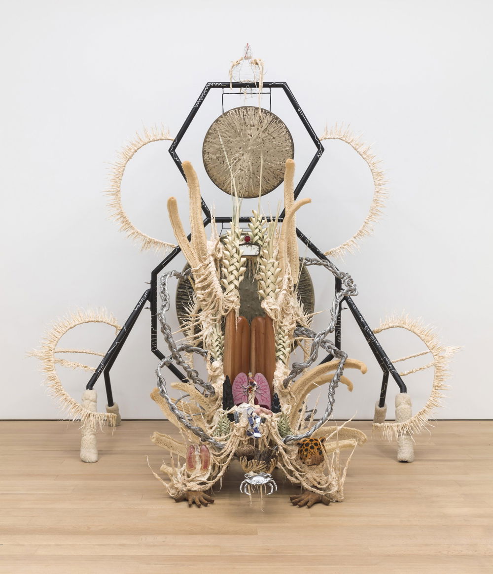 Image of shrinelike mixed-media sculpture in a white gallery space. A gong hangs from a hexagonal frame amidst woven circular frames and other appendages. Placed near the bottom is a medical model of lungs, two sculpted hands placed on the floor, and a crab centered between them.