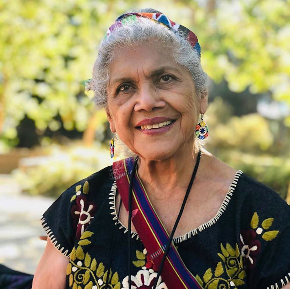 Ofelia, a Chicana elder woman with her white hair tied back with colorful Guatemalan textile headband, dons beaded huichol flower earrings, a black-corded medicine bag hanging from her neck, and a hand-loomed strap from a shoulder bag crosses her chest. She wears a black huipil with hand embroidered white flowers and olive green embroidered leaves. She looks up as she smiles, set in front of an out-of-focus hedge at Grand Park in Los Angeles Civic Center.