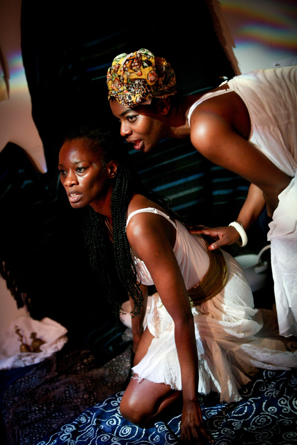 Two black women, both wearing white dresses, pose on a stage covered in colorful, patterned textiles. One woman leans over the other, who is kneeling, and seems to whisper in her ear.