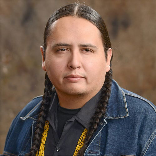 A headshot of a brown man with two brown braids on either side of his head. He looks directly at the viewer with a stoic expression. He is wearing a jean jacket and a yellow beaded necklace.