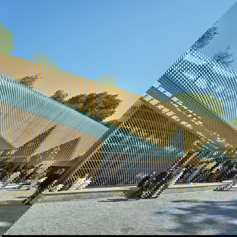 A timber and metal porch structure shelters a group of people from the sun.