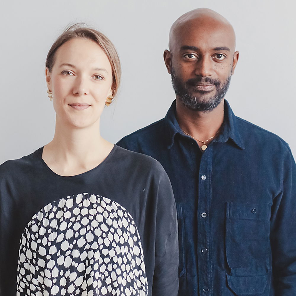 Two people stand side by side, facing us directly. Emanuel is a Black man with a shaved head and a neatly-trimmed beard wearing a navy corduroy shirt with black jeans. Jen is a white woman with light-brown hair wearing a long-sleeved black dress with a large white circle on the front.