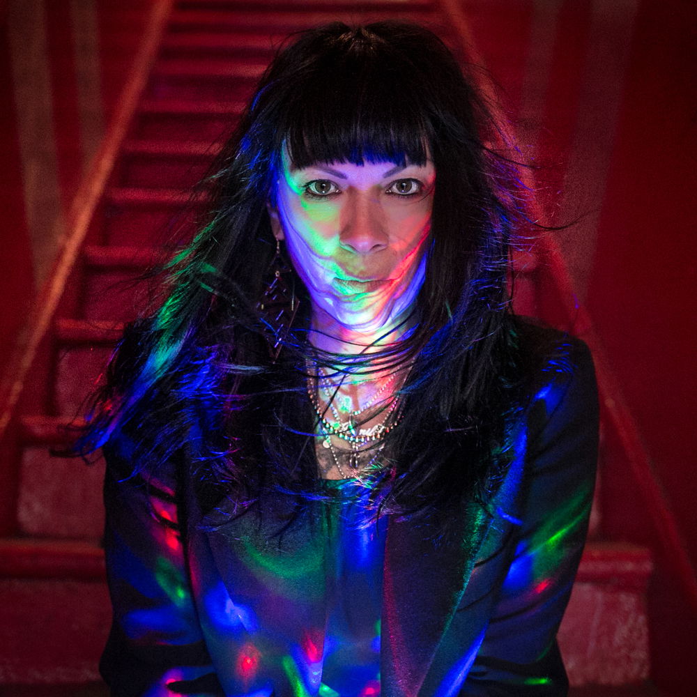 Jennifer, a woman with pale skin, long dark hair, and bangs, sits at the bottom of a narrow red staircase. Her hair blows around from an unseen source. She is looking at the camera with green, red and blue lights speckled across her face and torso. She is wearing a black jacket and no less than five necklaces.