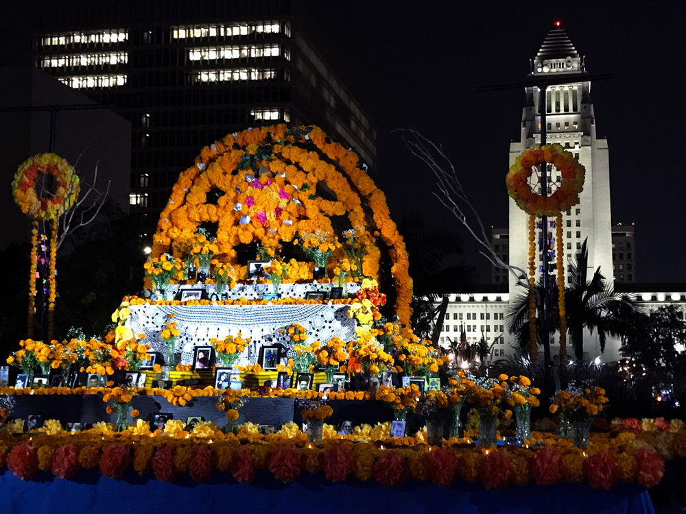 A multi-level Dia de Los Muertos altar covered with textiles, photographs of the recently departed, fresh marigold bouquets, hand crocheted tablecloth, and candles. Handmade paper flowers arranged in two wreaths frame the altar, and a giant arch with a sunburst motif forms its center.