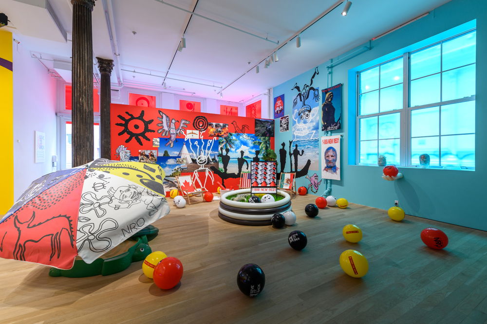 A gallery installation with objects placed around the floor. Lawn chairs are decorated with the Canadian, American, and Mexican flags. In the center, a black-and-white striped baby pool is filled with green grass and an alligator. Red, green, and yellow plastic balls have texts that read, "give it back." On the walls of the gallery hang stylized drawings and photographs of stereotypical portrayals of indigenous folks, oversaturated photographs of oceans, and posters advertising “1-888-NewRed1.”
