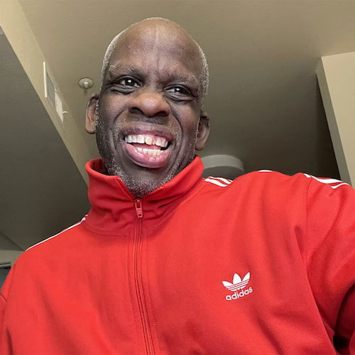 A portrait looking up onto the face and torso of a Black man, as if the camera were situated from slightly below. The man has short-cropped silver hair on his head and silver stubble on his face. He gives a big smile that is almost as vibrant as his red Adidas tracksuit.