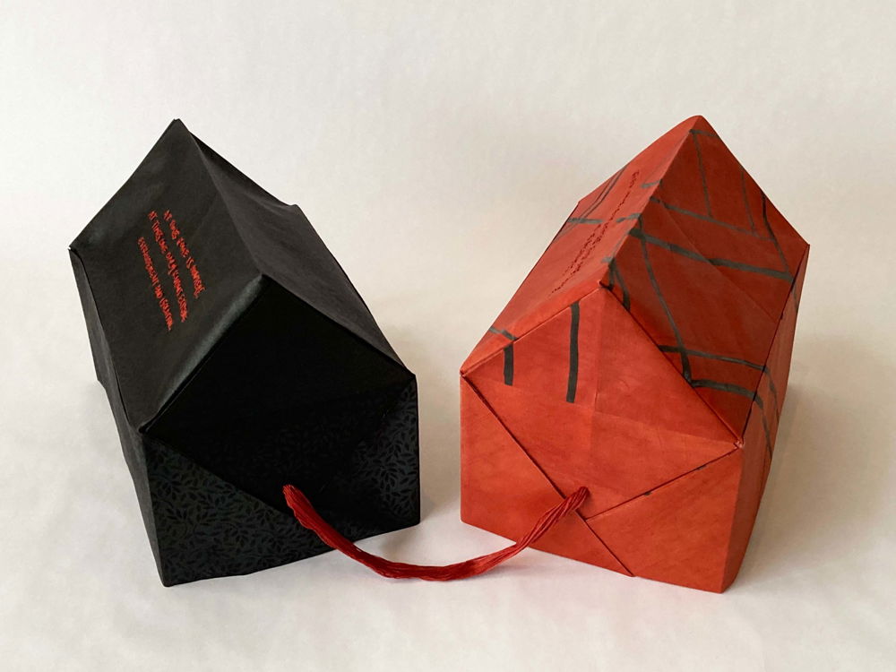 A photo of two house-like forms, one black and one red, folded origami-style from Nigerian head ties with a red strand connecting them. The house form on the left is patterned subtly with tiny oval leaves and red embroidered text, and the form on the right is covered in hand-drawn lines.