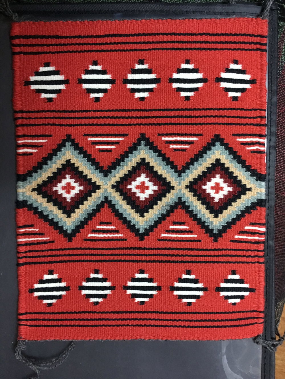 A blanket with a red background and three large diamonds in the center. The diamonds contain borders of teal, cream, and maroon lines radiating out from a red cross at the center of each shape. Smaller rows of smaller black and white diamonds border the top and bottom of the blanket.