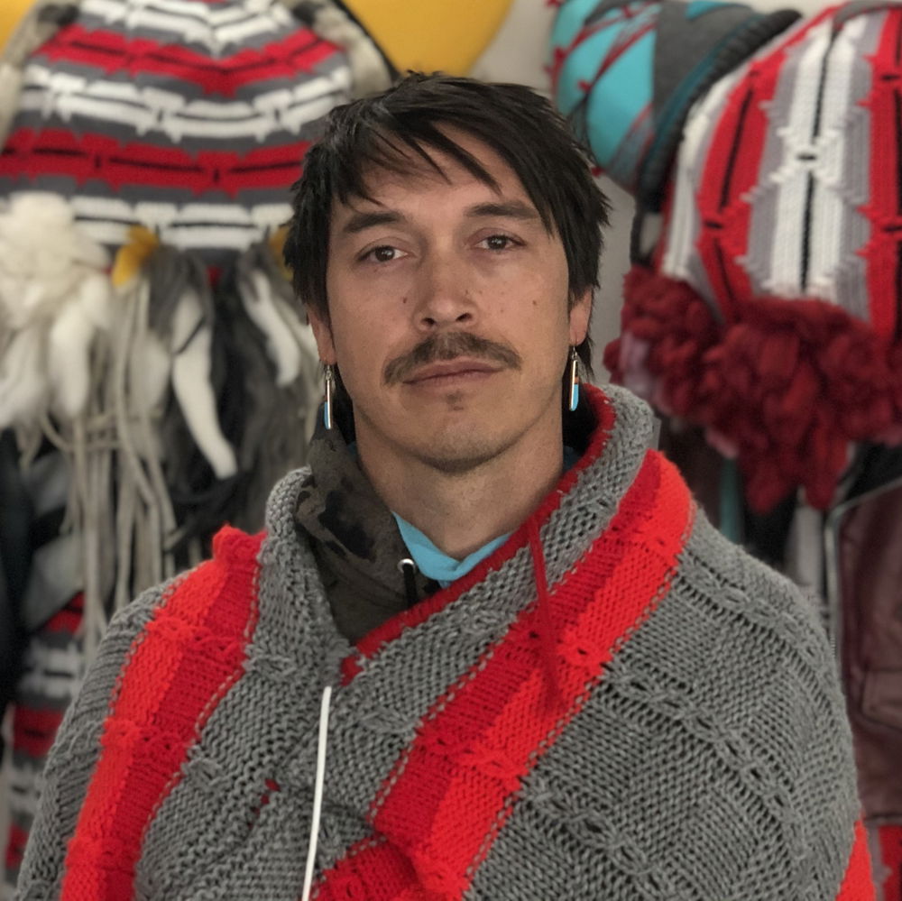 Cannupa, an Indigenous person of Mandan, Hidatsa, Arikara, Lakota heritage with brown hair, brown eyes, and a mustache, stands in their studio in front of two life-sized buffalo figures wearing regalia made of crochet, felt, ceramic, and steel. Cannupa is wearing a red and grey crochet cape with knife-shaped turquoise earrings.