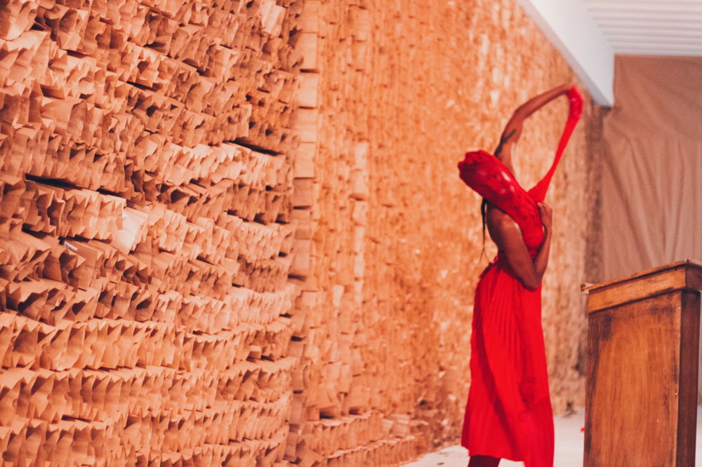 A person dances in a room where the walls have been lined with brown paper bags. They stand to the right of the frame, wearing a bright red dress covering their body and face. One outstretched hand pulls a length of the fabric upward.