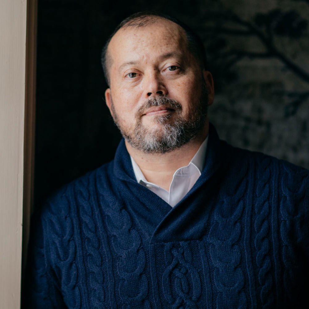 Alexander, a mixed race Korean American man of 53, is dressed in a navy cable-knit sweater and dress shirt. The wallpaper behind him is charcoal and black, and his face is lit by the sun coming from the window.