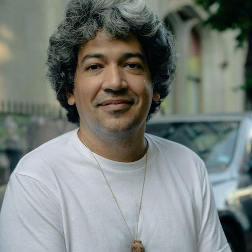 A portrait of a man sitting outside on a city block. The man has light brown skin and a full head of lustrous silver hair. He smiles at the viewer in a relaxed pose and wears a white t-shirt and a necklace.