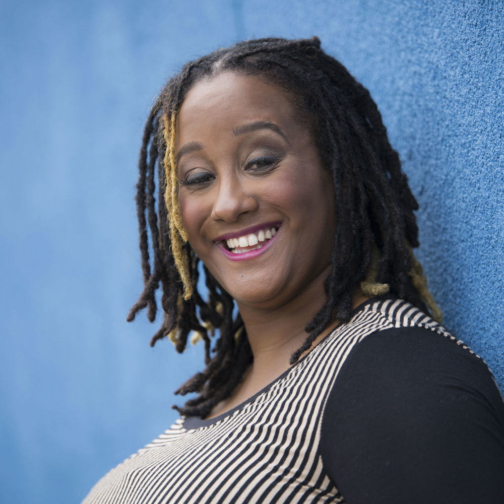 Kayla poses in profile in front of a light blue background. She is a milk chocolate-skinned Black woman who is wearing a long sleeve black and tan striped shirt. She has shoulder length dreadlocks and a glowing smile.