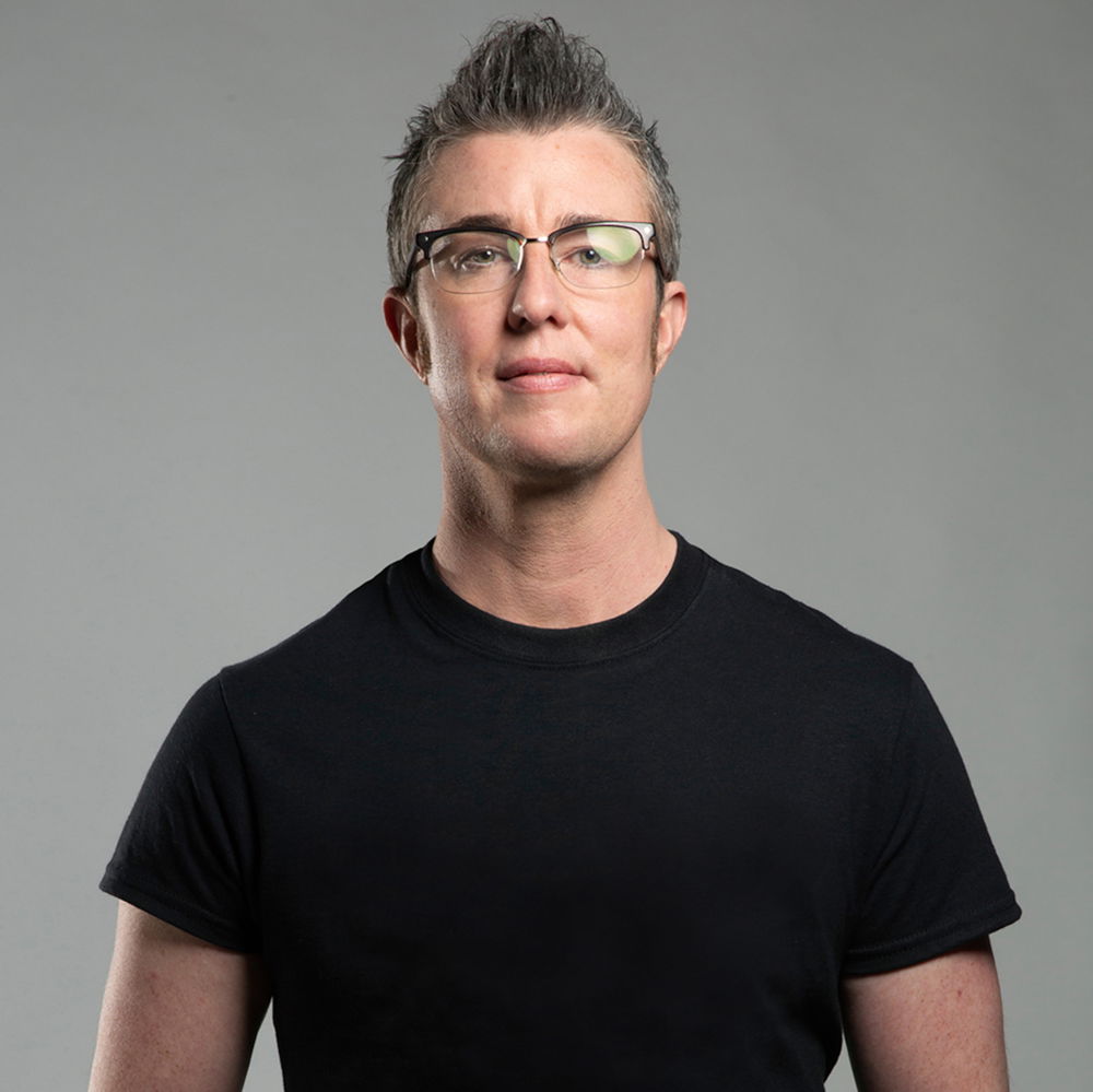 A light-skinned trans-masculine person with a warm and confident smile. He is wearing a black T-shirt, glasses, and has a short brown pompadour.