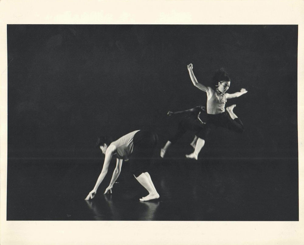 A black and white photograph of three dancers on a black stage. One bends over, touching the ground. To the right, a dancer flies, mid-jump, arms out, one leg bent behind her. The third dancer, arms and legs outstretched, is blurred in motion behind them.