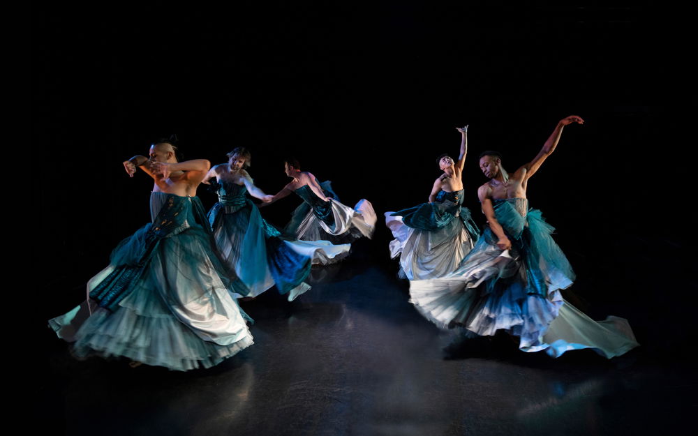 Five dancers twirl along a dark stage in teal gowns with sleeveless bodices. The dancers reach their arms up and out in different directions full of wistful yearning.