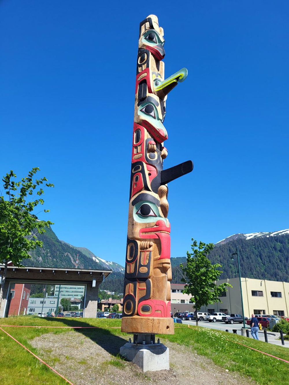 A tall totem pole in front of a mountain landscape. The structure is a long wooden pole ornately carved with three stylized animals one sitting on top of the other and painted in a limited color palette of red, black, green, white, and gray-blue.
