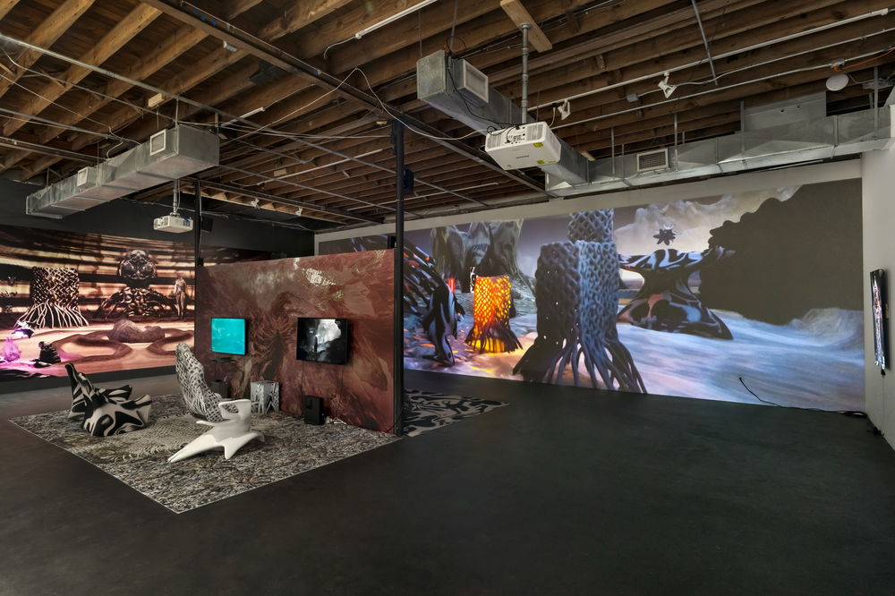 An installation view of a gallery show in a large room with tall ceilings: in the background is a wall-sized projection of a video game, depicting a surreal, abstracted landscape, and in the foreground is a temporary wall outfitted with an amorphous texture and television monitors featuring two playable game demos. Nearby are biomorphic textured carpets and angular interactive gaming chairs.