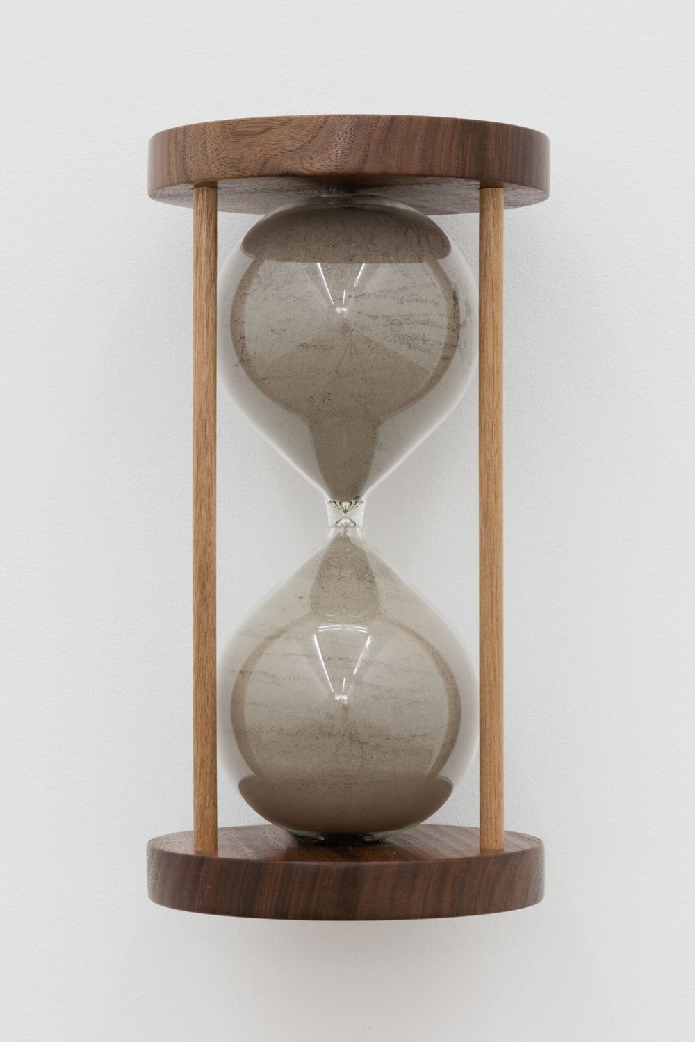An hourglass with both sides completely filled with gray dust is hung on the wall. The glass is set between a dark wooden base and cap with two visible wooden spindles. Inside the glass the gray dust forms cracks and layers.