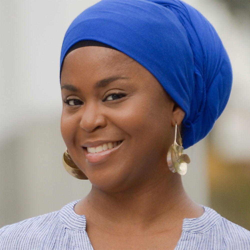 A Black woman smiles warmly into the camera. She wears a royal blue headwrap and large silver earring hoops.