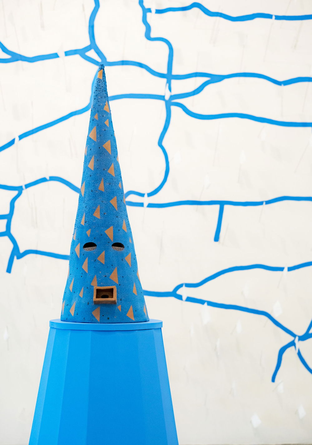 A ceramic cone sculpture that has its base resting on a medium blue pedestal. The sculpture has also been painted a medium blue with dark-blue dots and warm-brown triangles. Towards the base of the cone are two eye-shaped holes and a square-like mouth protruding outward.