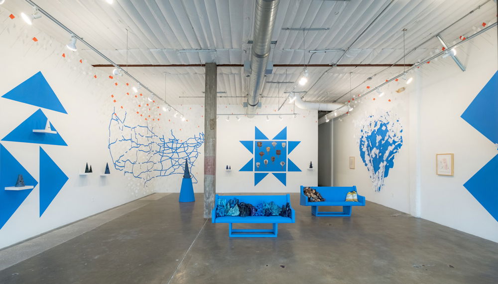 A gallery with white walls featuring a design of triangles, geometric shapes, an outline of the US, and an organic shape resembling an enormous puddle; all are painted in the same medium-blue color. Along the walls are shelves with cone-shaped sculptures and small orange-and-white flags. In the center of the space are two medium-blue benches covered with indecipherable objects.