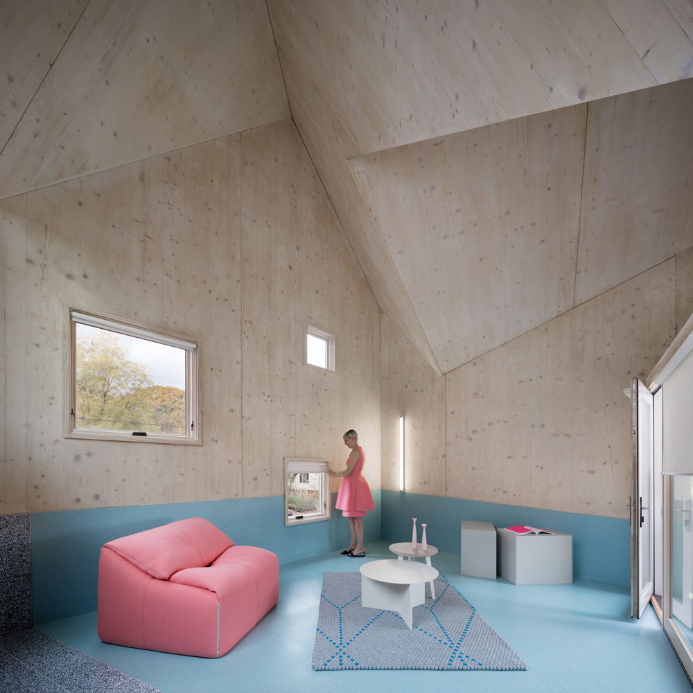 A home interior with a vaulted, jagged ceiling and powder blue flooring. All of the walls and roof surfaces have a soft wood finish. A model looks out of a window wearing a bubblegum pink dress that matches a plush loveseat nearby.