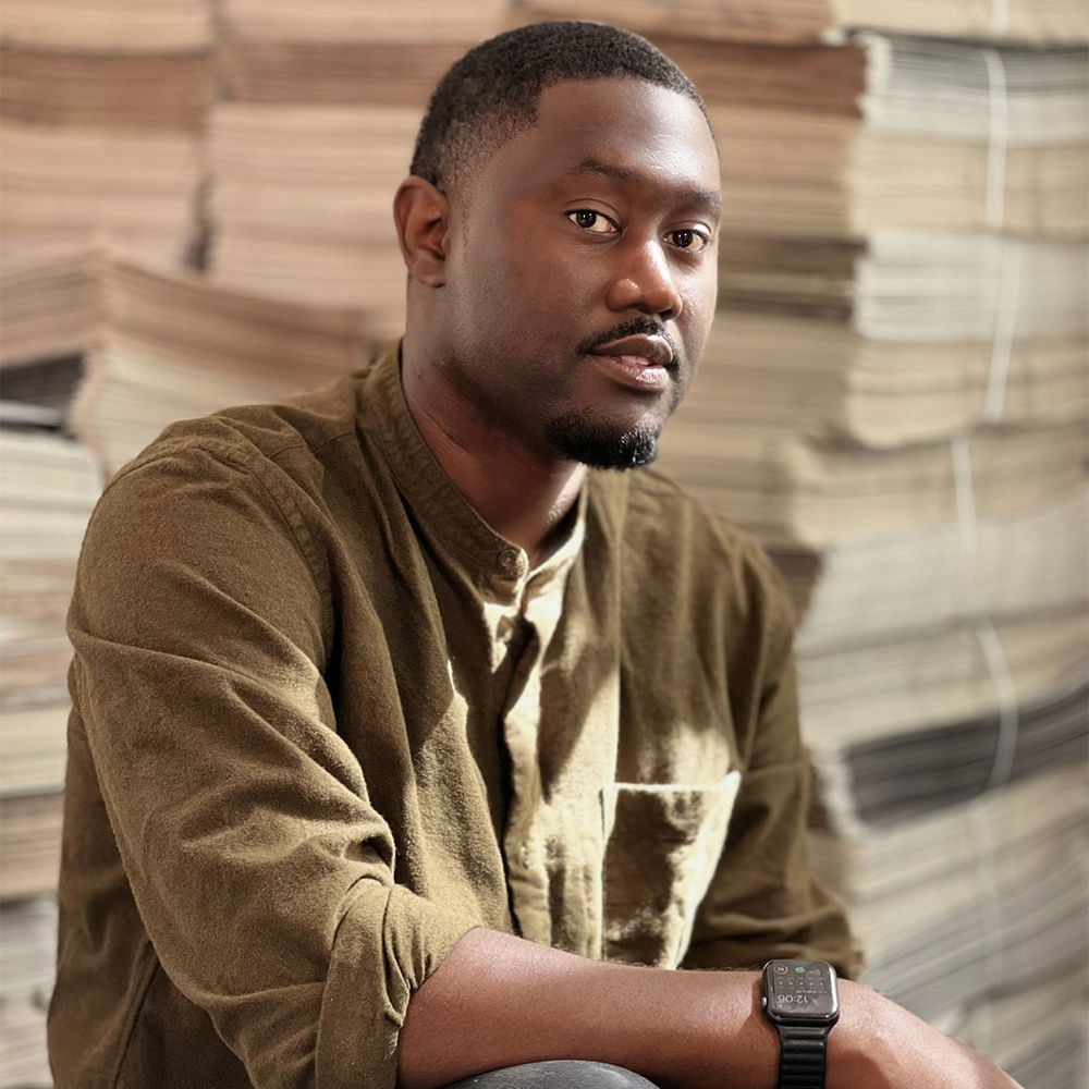 A Black man with a Caesar haircut sits in front of a stack of newspapers. He wears a green long-sleeved button-down shirt and denim jeans.