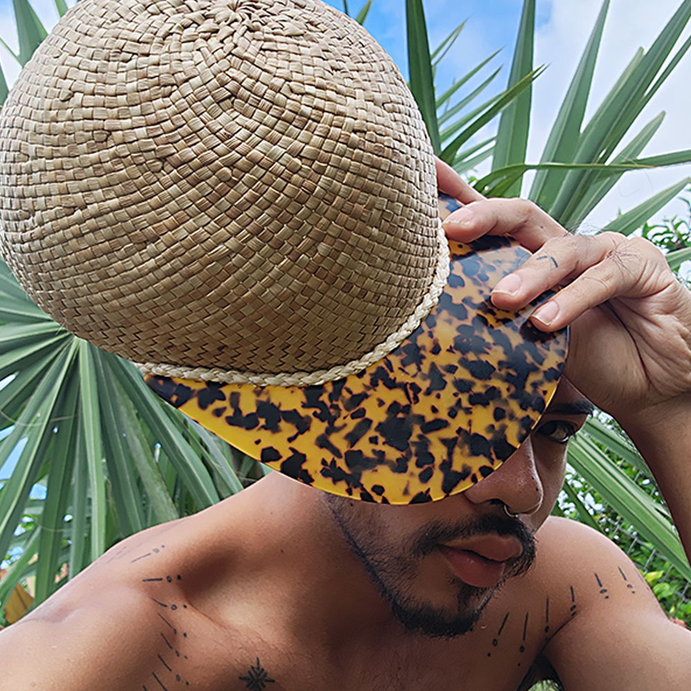 The artist holds up a woven cap to the camera, partially covering their face. The crown of the cap is woven tightly from pandanus. The bill is made from a material resembling tortoiseshell. A thin braid divides the bill from the crown.
