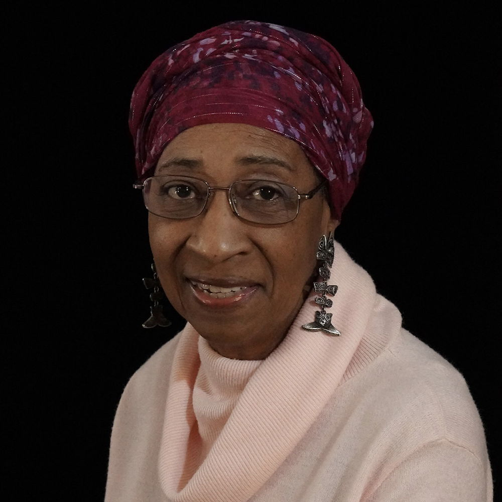 A slender woman with brown skin sits for her portrait, smiling slightly. She wears a light pink turtle-neck sweater, dangling earrings, and magenta head wrap.