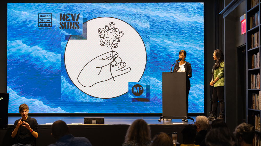 Two women stand towards the right of a stage behind a podium. A projected image fills a screen from the top to the bottom of the stage. The image is of a digital ocean-like background. In the center of this background is a white circle with a vector drawing of a hand holding a single flower. The petals of the flower are made of line drawings of eyes and ears.